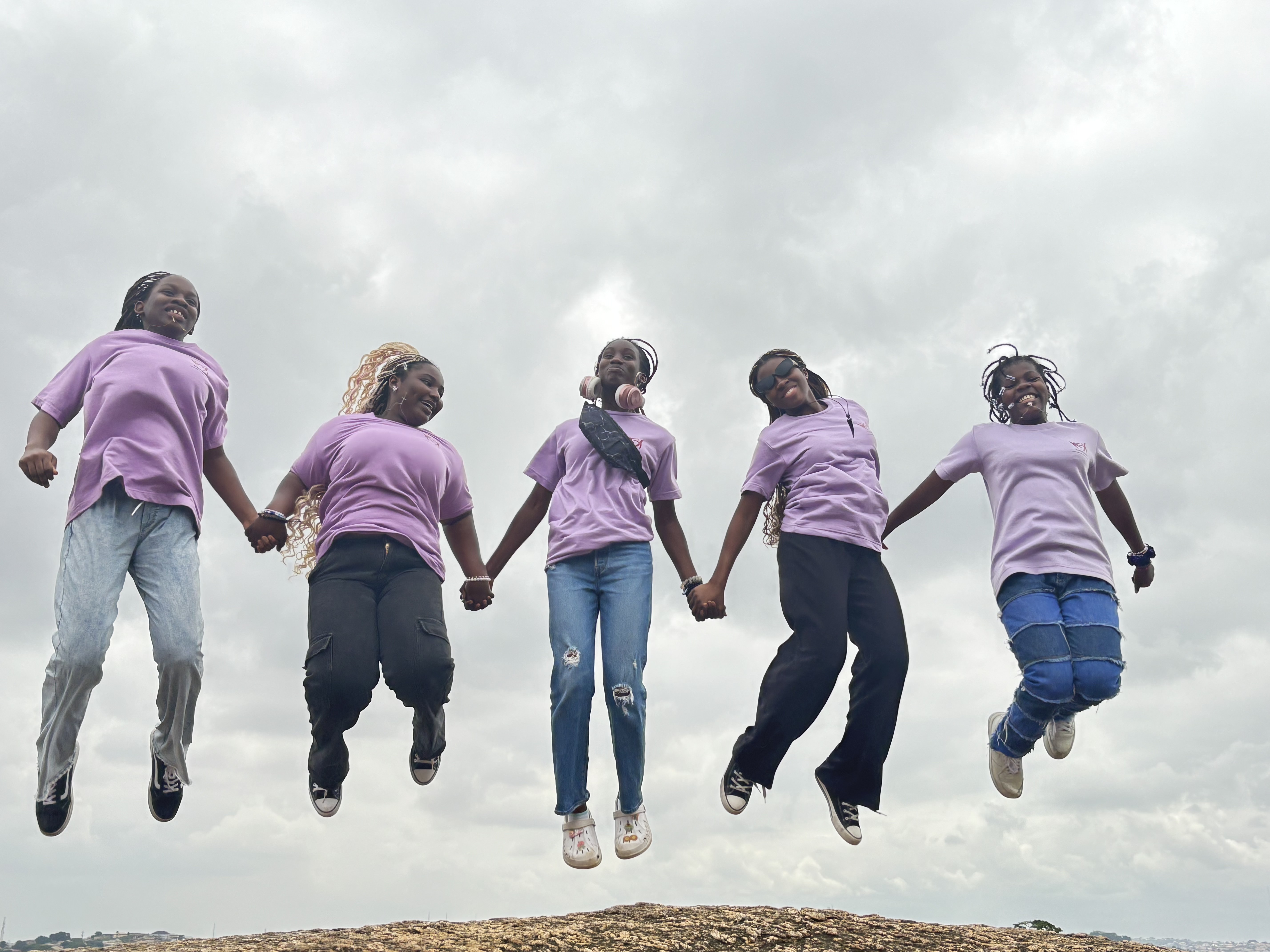 Five girls in purple t-shirts jumping joyfully in the air against a cloudy sky background