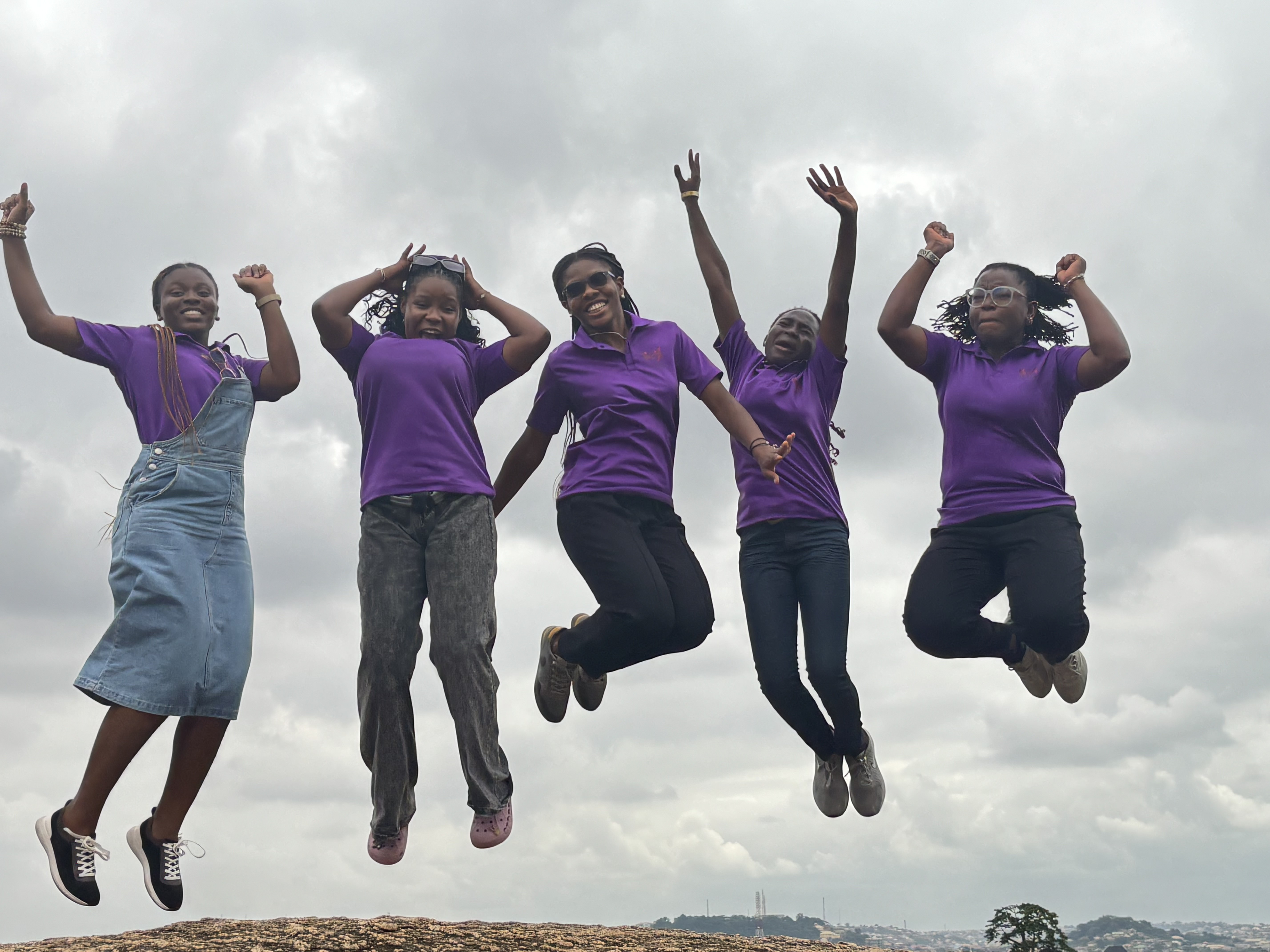 Five volunteers in purple Kamp Konnect shirts jumping joyfully in the air with city skyline in background, showing team celebration and energy